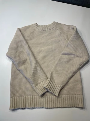 Beige stickad tröja  - En stilren beige stickad tröja med rund halsringning och ribbade muddar vid ärmslut och nederkant. Tröjan har en avslappnad passform och är perfekt för lager på lager. Materialet känns mjukt och bekvämt mot huden.