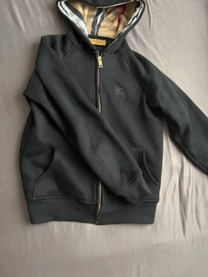 Svart hoodie från Burberry - Snygg svart hoodie från Burberry med dragkedja och klassiskt rutigt mönster i huvan. Tillverkad i mjukt bomullsmaterial och har två fickor framtill. Perfekt för en chill och stilren look.