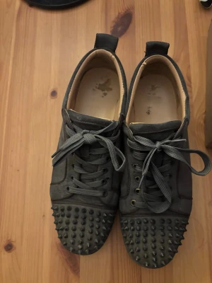  Christian Louboutin med spikar  - Unika mörkgrå sneakers från Christian Louboutin med matta nitar över tån och klassiska röda sulor. Skorna är låga och har snörning samt ovandel i mocka. Perfekta för dig som vill sticka ut med en edgy och exklusiv stil.
