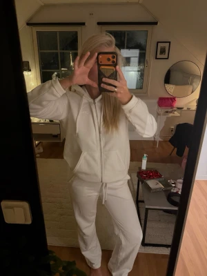 Vit mjukisdräkt från J.Lindeberg - Säljer en vit mjukisdräkt från J.Lindeberg med dragkedja, huva och fickor på tröjan samt matchande byxor med dragsko i midjan. Perfekt för chill dagar hemma. Materialet är mjukt och skönt, troligen bomullsmix. Enkel och stilren design med liten logga på bröstet samt på låret. Byxan är en rak modell i storlek xs och tröja i L. Köpt varje del för ca 1000kr/st. 