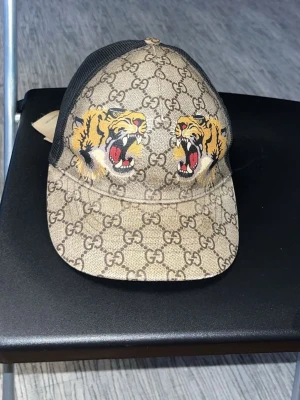 Gucci keps med tigertryck - Snygg keps från Gucci med klassiskt GG-mönster i beige och brunt. Framtill pryds kepsen av två färgstarka tigerhuvuden. Baksidan är i svart mesh för extra ventilation. Den kommer med box kvitto och allt som finns när man köper den på butik, den är äkta! Pris kan diskuteras!
