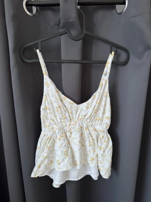 Blommig vit linnetopp med volang - Söt vit linnetopp med små gula blommor och justerbara axelband. Toppen har en volangig nederdel och smock i ryggen. En perfekt sommartopp💛