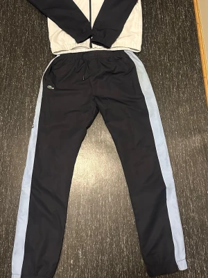 Lacoste tracksuit  - Sportigt set från Lacoste med jacka och byxor i marinblått, vitt och ljusblått. Användt sparsamt. Inga slitningar. 