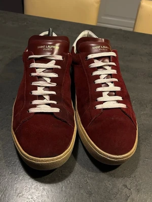 Vinröda Saint Laurent sneakers i mocka - Snygga vinröda sneakers från Saint Laurent i mjuk mocka med vita skosnören och beige sula. Finns en lite defekt som ni ser på bild 5 längst fram på skon annars inga defekter, ingen box medföljer 