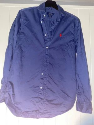Blå skjorta från Polo Ralph Lauren - Klassisk blå skjorta från Polo Ralph Lauren med lång ärm och knappar framtill. Skjortan har den ikoniska röda logotypen broderad på bröstet och är tillverkad i bomull. Perfekt för en stilren och avslappnad look. Använt max 3 gånger