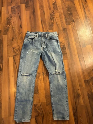 Ljusblå slitna raka jeans från Levis  - Säljer ett par ljusblå jeans med raka ben och slitningar på både lår och knän. Jeansen har klassisk femficksmodell och knappgylf. Perfekta för en avslappnad och trendig look.