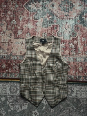 Rutig väst från H&M med bälte - Snygg rutig väst från H&M i beige, brunt och svart med klassisk knäppning fram och justerbart bälte bak. Västen har spetsig nederkant och är helfodrad med ljus insida.