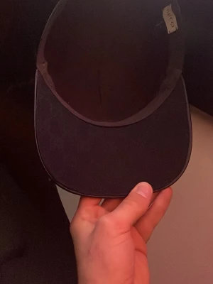 Svart keps från Gucci med röd/grön detalj - Boxen kommer med G kepsen! Säljer en svart keps från Gucci med klassisk böjd skärm och den ikoniska rödgröna randen på sidan. Kepsen har diskret Gucci-mönster på skärmen och är tillverkad i ett slitstarkt tygmaterial. Perfekt för dig som vill ha en stilren accessoar med lyxig känsla.