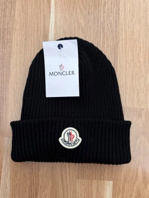 Svart ribbstickad mössa från Moncler - Snygg svart ribbstickad mössa från Moncler med uppvikt kant och klassisk Moncler-logga framtill. Tillverkad i mjukt material som håller dig varm och ger en clean look. Perfekt accessoar för dig som vill ha både stil och komfort. Priset är fast:)