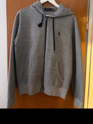 Grå hoodie från Polo Ralph Lauren - Snygg grå hoodie från Polo Ralph Lauren med dragkedja, svart logga broderad på bröstet och två fickor framtill. Huva med svarta snören och mjukt material som känns skönt mot huden. Perfekt för en avslappnad och stilren look.