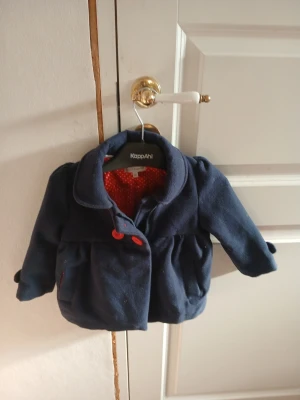 Mörkblå kappa från KappAhl barn - Söt mörkblå kappa för barn från KappAhl med röda knappar och rödprickigt foder. Kappan har klassisk krage, två fickor fram och dekorativa detaljer vid ärmsluten. Perfekt för kyligare dagar och ger en stilren look.