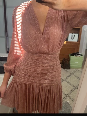 MAJE Glittrig kort klänning i roséguld  - Supersnygg kort klänning i glittrigt roséguld med v-ringning och långa ärmar. Klänningen har markerad midja, plisserad kjol och lätt puffade ärmar. Perfekt för dig som vill sticka ut med en trendig look. 34/XS only one time used 🩷🩷