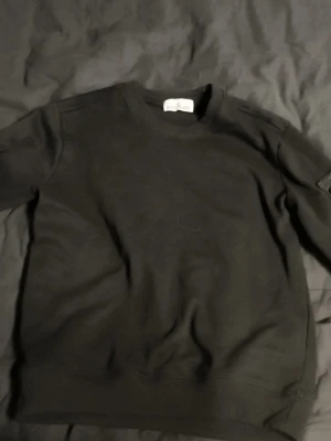 Svart sweatshirt från Stone Island - Säljer en svart sweatshirt från Stone Island med klassisk rund hals och ribbade muddar. Tröjan har en diskret patch på ärmen och är tillverkad i mjukt bomullsmaterial. Perfekt för en clean och stilren look. Pris går att diskutera 