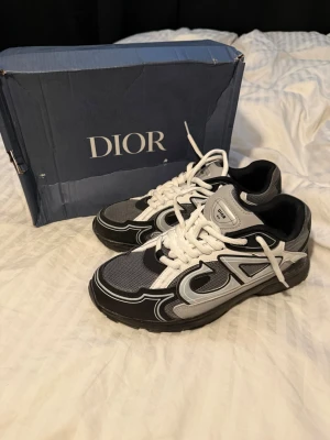 Dior grå och svarta sneakers - Snygga Dior sneakers i svart, grått och vitt med coola detaljer och vita snören. Skorna har en sportig siluett med mesh och läderpaneler samt tydlig Dior-logga på plösen och innersulan. Perfekta för dig som vill sticka ut med exklusiva sneakers.