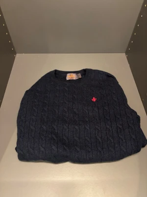 Mörkblå kabelstickad tröja från  Morris - Kabelstickad mörkblå tröja från Morris i 100% merinoull. Tröjan har rund halsringning och en liten röd broderad logga på bröstet. Perfekt för dig som gillar klassisk och stilren design med en twist.