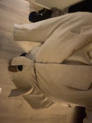 Beige kappa med knytskärp - Stilren beige kappa med bred krage och knytskärp i midjan. Kappan har en rak passform och långa ärmar, perfekt för lager på lager. Materialet ser ut att vara mjukt och lite tjockare, vilket gör den skön under kyligare dagar.