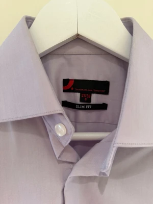 Ljuslila slim fit skjorta Dressmann - Säljer en stilren ljuslila skjorta från Dressmann i slim fit-modell. Skjortan har klassisk krage, knappar framtill och är perfekt för dig som gillar en smalare passform. Enkel och clean look som funkar till många olika outfits.