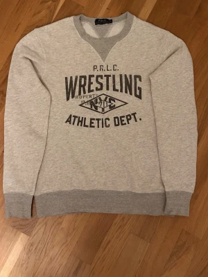 Grå sweatshirt från Polo Ralph Lauren storlek S - Säljer en ljusgrå sweatshirt från Polo Ralph Lauren med tryck i svart text 'Wrestling Athletic Dept.' på bröstet. Tröjan har ribbade muddar vid ärmslut och nederkant samt en V-detalj vid halsen. Perfekt för en sportig och avslappnad stil.