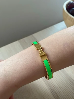 Grönt och guldigt armband H - Snyggt armband från H i guldmetall med gröna emaljdetaljer. Armbandet har den ikoniska H-formade låset och en stilren, rundad design som känns både lyxig och modern. Perfekt accessoar för att lyfta din outfit med en färgklick.