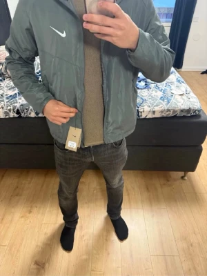 Grå vindjacka från Nike med huva - Snygg grå vindjacka från Nike med vit logga på bröstet och praktisk huva. Jackan har dragkedja framtill och två sidofickor. Tillverkad i lätt och vindtätt material, perfekt för blåsiga dagar. Enkel och stilren design som passar till det mesta.