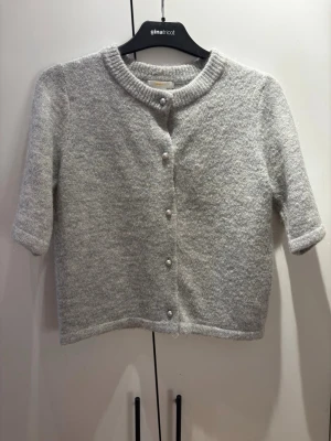 Grå kofta med pärlknappar Gina Tricot - Mysig grå kofta från Gina Tricot med korta ärmar och rund hals. Koftan har pärlknappar framtill och är stickad i ett mjukt material som ger en fluffig känsla. Perfekt för lager på lager och enkel att matcha med olika outfits.