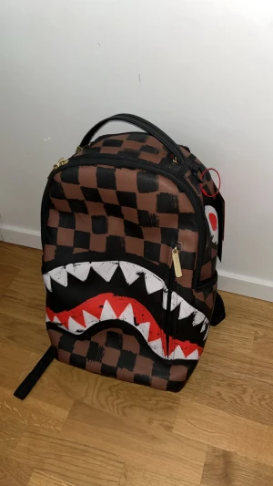 Sprayground brun ryggsäck med tänder - Limiterad,Perfekt skick och väldigt effektivt på grund av att de är jätte många fak. PRISET ÄR INTE HUGGET I STEN. ryggsäck från Sprayground i brunt och svart rutmönster med ikoniskt hajtandsmotiv. TAR BYTEN.
