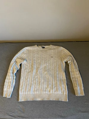 Ljusgrå kabelstickad tröja Gant - Kabelstickad tröja i ljusgrått från Gant med rund halsringning och diskret logga broderad på bröstet. Tröjan har långa ärmar och ribbade muddar vid ärmslut och nederkant. Perfekt för en stilren och klassisk look. Tröjan är i listad i model 170cm/15 år, sitter som M. Pris kan såklart diskuteras!