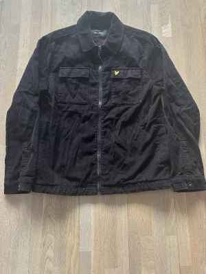 Svart overshirt från Lyle & Scott - Stilren svart overshirt från Lyle & Scott i manchestertyg. Två bröstfickor med lock, dragkedja framtill och klassisk gul logga på vänster bröst. Perfekt lager-på-lager-plagg med avslappnad passform och skjortkrage.