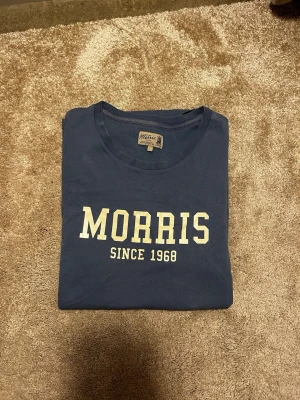 Blå t-shirt från Morris - Snygg blå t-shirt från Morris med vit text 'MORRIS SINCE 1968' på bröstet. Klassisk rund halsringning och normal passform. Mycket bra skick. Priset ej hugget i sten. Hör av er vid frågor eller funderingar! 
