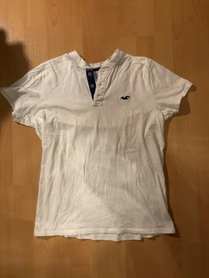 Vit t-shirt/henley shirt Hollister - Vit t-shirt/henley shirt från Hollister med korta ärmar och blå detaljer vid halsringningen. T-shirten har knappslå med två träknappar och Hollisters logga broderad på bröstet. Tillverkad i mjuk bomull för en skön känsla.