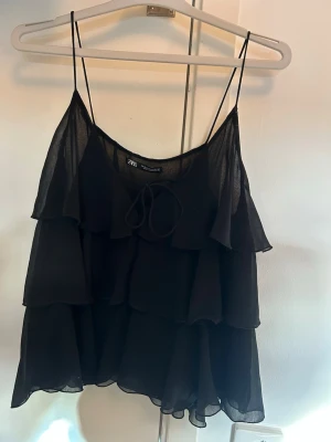 Svart volanglinnetopp från Zara - Säljer en svart linnetopp från Zara med tunna axelband och flera lager volanger. Toppen är luftig och har en lätt transparent känsla, perfekt för varma dagar. Snygg detalj med knyt framtill som ger en extra touch.