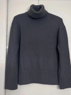 Mörkgrå polotröja i stickat - Säljer en mörkgrå stickad polotröja med bred ribbstickning vid ärmslut och nederkant. Tröjan har en hög, mysig polokrage och rak passform. Perfekt för kalla dagar och enkel att matcha med jeans eller kjol.