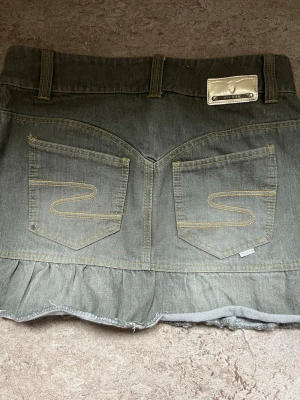 ”Grå” jeanskjol med volang  - Säljer en grå jeanskjol  med volangkant nedtill och snygga bakfickor med broderade detaljer. Kjolen har bälteshällor, knapp och dragkedja framtill samt en metallbricka med logga bak. Perfekt för en trendig och avslappnad look.