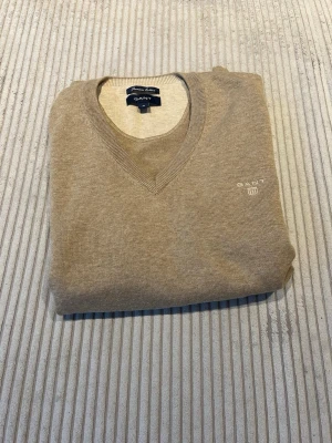 Beige V-ringad tröja från Gant - Storlek:M, normal i storleken.                                 Klassisk beige V-ringad tröja från Gant med diskret logga broderad på bröstet. Tröjan har långa ärmar, ribbade muddar och förstärkta armbågslappar i matchande färg. Tillverkad i mjukt material som känns skönt mot huden.