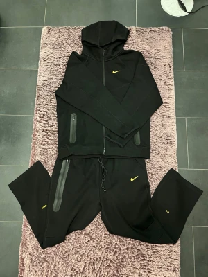Svart Nike nocta hoodie och byxa med gul logga - Snygg svart hoodie från Nike med dragkedja och huva med snygga svarta byxor. Har gul Nike-logga på bröstet och detaljer i gult på ärmen. Två fickor med dragkedja framtill. Har gul Nike logga på vänster benen och gul design runt om byxan. Har två fickor en med dragkedja i höger benet och en inte i vänstra benet. Tillverkad i mjukt och bekvämt material, perfekt för en sportig och avslappnad stil. Är XL men passar som L. Aldrig använt!!