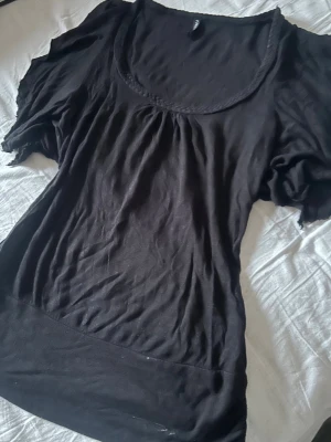 Svart topp med puffärmar - Svart topp med rundad halsringning och puffiga kortärmar. Tunn och mjuk i materialet, perfekt till jeans eller kjol. Dekorativ kant runt halsen ger en extra detalj. Enkel och stilren design.