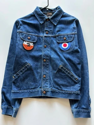 Blå jeansjacka med patches - Jeansjacka med två bröstfickor som har knäppning och . Jackan har även två snedställda fickor framtill. Knäppning med knappar framtill och vid ärmslut.  📌 Detaljer Färg – Blå Storlek – 140 Skick – Begagnad  📏 Mått Axel till axel – 42 cm Axel till ned