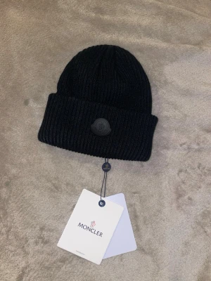 Svart ribbstickad mössa Moncler - Säljer en svart ribbstickad mössa från Moncler med uppvikt kant och diskret svart Moncler-logga framtill. Mössan är tillverkad i ett mjukt och varmt material som passar perfekt för kyliga dagar. Klassisk och stilren design som funkar till det mesta.