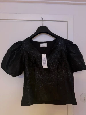 Glittriga blus från neo noir - Snygg svart topp från Neo noir med glittrig finish och puffiga korta ärmar. Toppen har v-ringning och croppad passform, perfekt för att sticka ut. Materialet känns lite styvt och skimrande. Nypris 799 kr