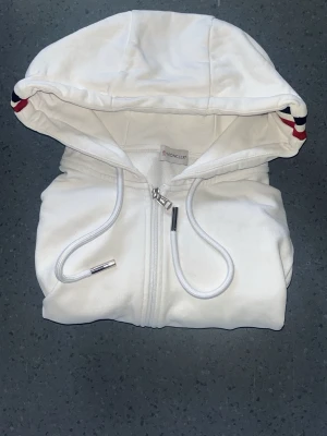Vit hoodie med dragkedja från Moncler - Snygg vit hoodie från Moncler med dragkedja framtill och två fickor. Huva med dragsko och coola rödblå detaljer på kanten. Tillverkad i mjukt material som känns skönt mot huden. Moncler-logga på ärmen för extra stilpoäng. Storleken är L men passar L