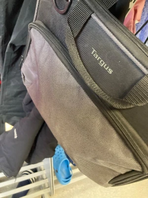 Svart datorväska från Targus - Snygg svart datorväska från Targus med flera fack och robust axelrem. Tillverkad i slitstarkt tygmaterial och har dragkedjor för säker förvaring av laptop och tillbehör. Perfekt för dig som vill bära din dator på ett stilrent sätt.