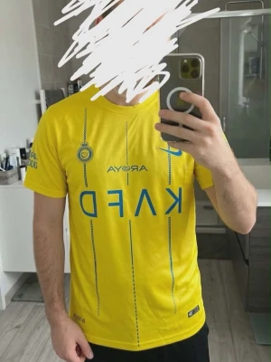 Al Nassr fotbollströja Ronaldo - Säljer en gul fotbollströja från Al Nassr med blå detaljer och tryck. På ryggen står det 'Ronaldo' och nummer 7. Tröjan har korta ärmar, rund hals och är tillverkad i ett lätt, andningsbart material som passar perfekt till träning eller match.