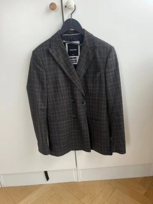 Rutig dubbelknäppt kostym från Jack & Jones - Snygg rutig kavaj med tillhörande byxor från Jack & Jones i mörkbrunt och grått med diskreta röda linjer. Dubbelknäppt modell med breda slag och två framfickor. Perfekt för en stilren och trendig look. Materialet känns mjukt och följsamt.
