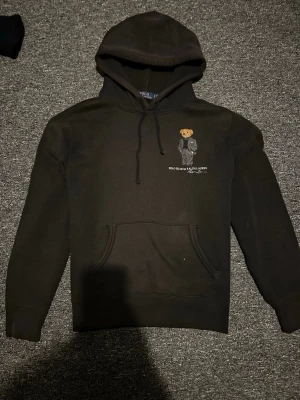 Svart hoodie Polo Bear Ralph Lauren - Svart hoodie från Ralph Lauren med Polo Bear-broderi på bröstet. Klassisk känguruficka framtill och justerbar huva med snören. Mjuk bomullskänsla och stilren design med ikonisk björn i kostym. Perfekt för dig som gillar streetwear och snygga detaljer. Äkta såklart jättebra kvalite speciellt vid björnen
