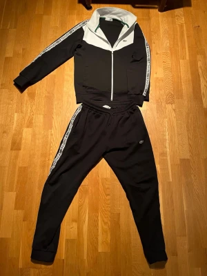 Svart och vit Lacoste tracksuit - Säljer en svart och vit tracksuit från Lacoste med dragkedja framtill och klassisk krage. Setet har vita detaljer och logga på både jacka och byxor samt vita ränder med Lacoste-tryck längs ärmar och ben. Perfekt för chill dagar hemma eller när du vill vara bekväm.