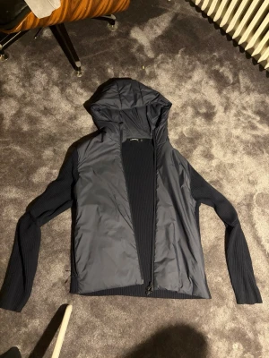 Mörkblå vindjacka med huva - Snygg mörkblå vindjacka med huva och dragkedja framtill. Jackan har stickade ärmar och sidor i ribbat material, medan fram- och baksidan är i slätt, vindtätt tyg. Perfekt för dig som vill ha en sportig och modern look.