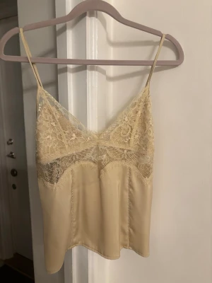 Beige spetslinnetopp med smala band Zara  - Säljer en beige linnetopp med tunna axelband och v-ringning. Toppen har fina spetsdetaljer över bysten och är tillverkad i ett glansigt, mjukt material som ger en lyxig känsla. Perfekt att styla lager på lager eller som den är.