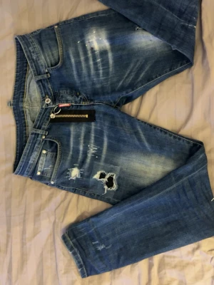 Blå slitna jeans från Dsquared2 - Säljer ett par blå jeans från Dsquared2 med slitningar, hål och coola detaljer. Jeansen har en stor silverfärgad dragkedja framtill och tydliga gula sömmar. Materialet är klassisk denim i bomull och passformen är smal. Perfekta för dig som gillar streetstyle och edgy look.