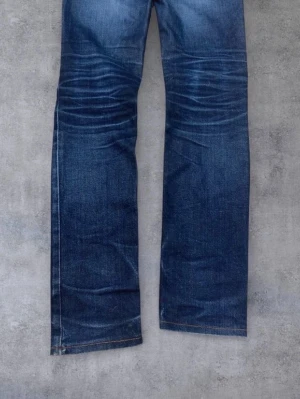 Blå raka jeans från Acne Studios - Säljer ett par klassiska blå jeans från Acne Studios med raka ben och snygga slitningar. Jeansen har orange kontrastsömmar och silverfärgad knapp med logga. Perfekta för en avslappnad och trendig look.
