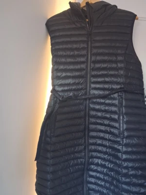 Svart lång dunväst med huva - Snygg svart lång dunväst NS med huva och dragkedja framtill. Västen har quiltad design, två fickor med dragkedja och ett midjeband för extra passform. Perfekt för lager-på-lager och streetstyle. Materialet är glansigt och lätt, typiskt för dunvästar.